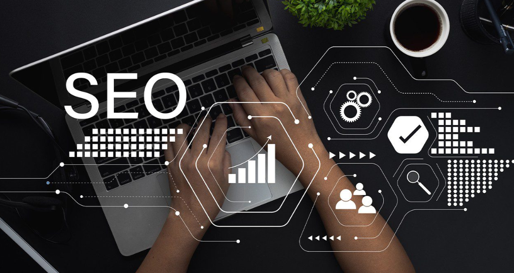 Uma visão aérea de mãos digitando em um laptop, com a palavra "SEO" em destaque no canto superior esquerdo da tela. Redes de linhas pontilhadas e ícones digitais como engrenagens, gráficos de barras ascendentes, um sinal de "check", pessoas e uma lupa cercam as mãos e o laptop, representando a conectividade e as diversas funcionalidades das ferramentas de SEO. Há também uma planta e uma xícara de café na mesa.