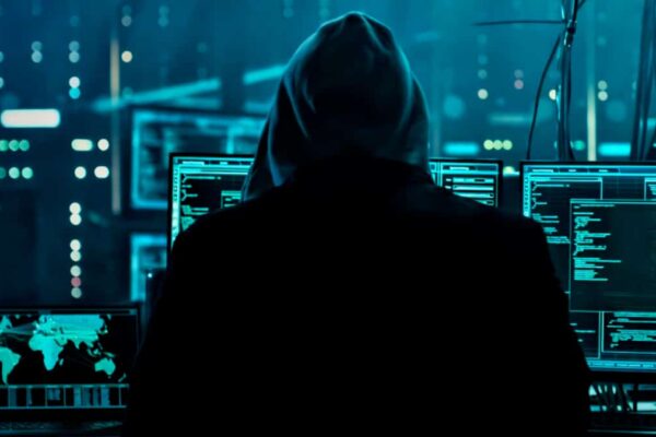 A imagem mostra a visão de costas de uma pessoa com um capuz, sentada em frente a uma série de monitores. A pessoa, que se assemelha a um hacker, está no centro da cena, em silhueta escura. Os monitores ao seu redor exibem códigos e gráficos digitais em tons de ciano e azul. Um dos monitores mostra um mapa-múndi, sugerindo a natureza global da atividade. O ambiente escuro e a iluminação digital no fundo criam uma atmosfera de espionagem e crime cibernético.