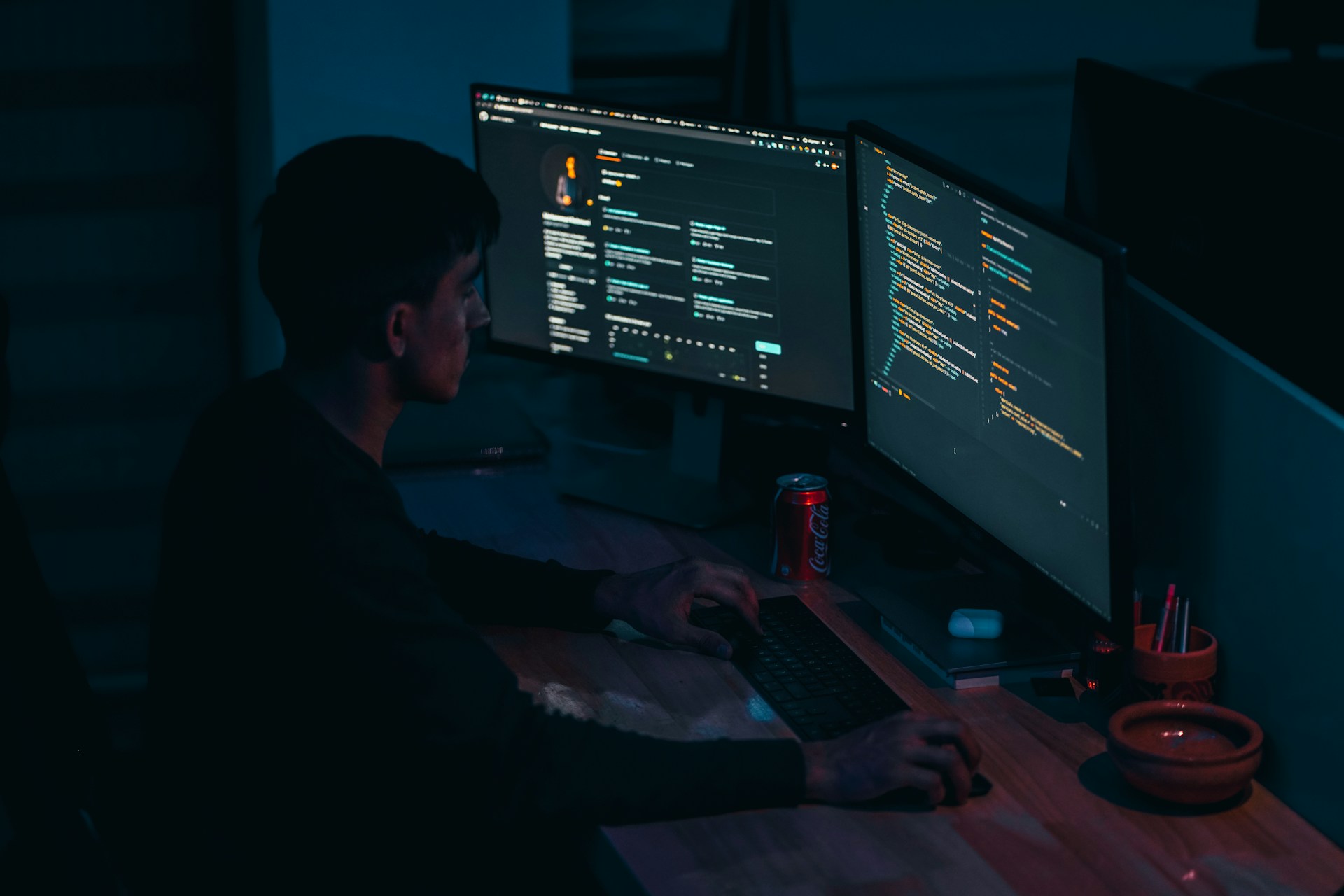 Jovem programador trabalhando em ambiente escuro com dois monitores exibindo códigos e interfaces de desenvolvimento, representando o universo das Linguagens de Programação e da tecnologia moderna.