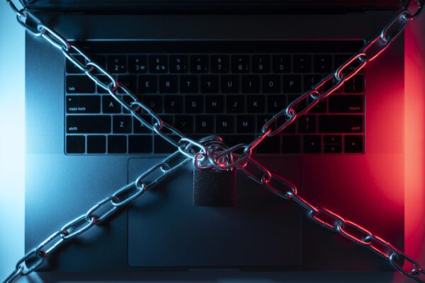 Um laptop preto visto de cima, com a tampa aberta revelando o teclado. Duas correntes de metal grossas se cruzam sobre o teclado e se prendem a um cadeado no centro. O laptop está iluminado com luzes de neon em azul e vermelho, criando uma atmosfera de urgência. A imagem simboliza a proteção da superfície de ataque de um dispositivo, representando a segurança e as medidas de proteção contra ameaças cibernéticas.