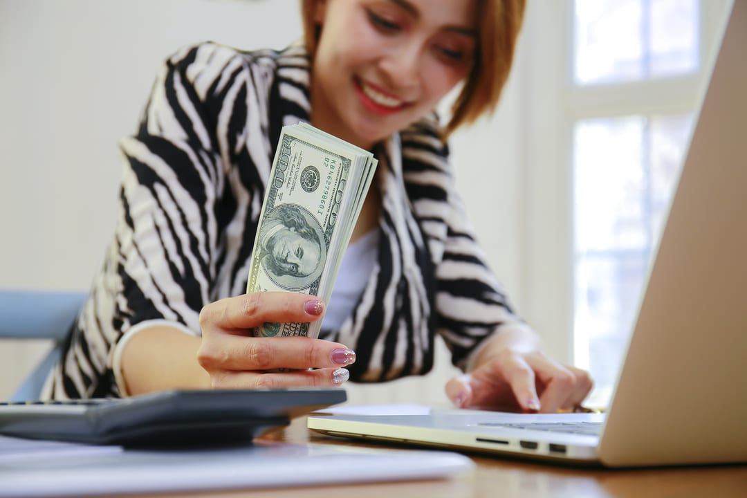 Uma mulher sorridente, vestindo um blazer com estampa de zebra, está segurando um maço de notas de 100 dólares em uma mão, enquanto a outra mão está no teclado de um notebook. A imagem, que é brilhante e iluminada, sugere sucesso financeiro ou o ato de ganhar dinheiro. A cena pode ser interpretada como a pessoa lucrando ou monetizando o conteúdo e a produtividade gerada por ferramentas como o Chat GPT. Uma calculadora está em primeiro plano na mesa.