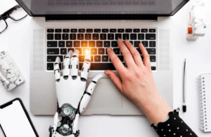 Uma visão de cima de um laptop onde uma mão humana e uma mão robótica estão digitando no teclado, com foco de luz na área das teclas. Representa a interação direta entre humanos e sistemas de IA/robótica na inserção de prompts.