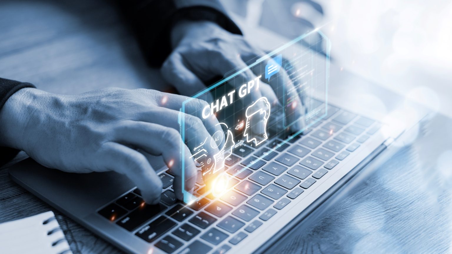 Close-up em tons de cinza e azul de mãos digitando rapidamente em um teclado de notebook. Um efeito de sobreposição digital futurista e luminoso, em tom ciano, paira sobre o teclado. Neste display virtual, a sigla "CHAT GPT" se destaca, acompanhada de ícones que representam comunicação, inteligência artificial e processamento de dados, simbolizando a interação entre humano e máquina ou a utilização de um chatbot de IA. A imagem sugere a aplicação da tecnologia de IA no trabalho ou na comunicação.