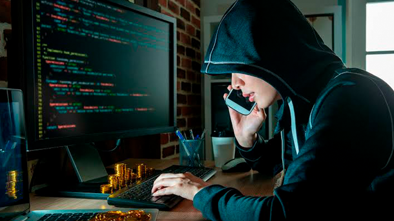 Pessoa vestindo um moletom com capuz (hacker) digitando em um teclado e falando ao telefone, com código de programação em uma tela de computador. A invasão de privacidade é real e o primeiro passo é tapar sua webcam.