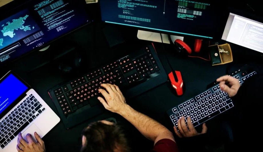Visão de cima de uma mesa escura com um setup de computador que inclui vários monitores exibindo códigos e mapas mundiais. Um profissional usa múltiplos teclados e mouses, enfatizando o ambiente técnico necessário para quem quer ganhar dinheiro hackeando ou programando.