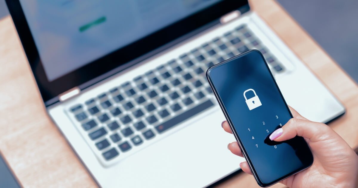 Uma mão segura um smartphone com uma tela de bloqueio (cadeado e teclado numérico) sobre um notebook aberto, representando segurança de dados. O contexto é importante, assim como tapar sua webcam.